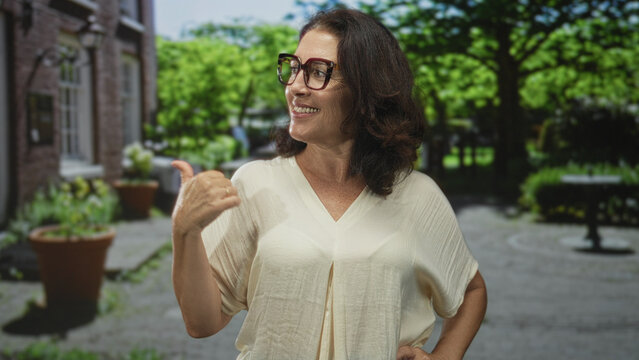 Woman smiling points thumb over shoulder toward building courtyard with potted plants and brick wall, wearing glasses and blouse; confidence friendly invitation.