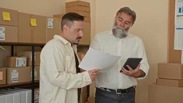 Two men hold paperwork and review parcels, one points to tablet while comparing shipping labels inside building; focused teamwork.
