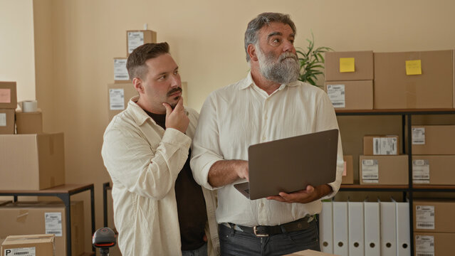 Two men with laptop, one hand on chin and one pointing at screen among stacked parcels in a storage building; teamwork.
