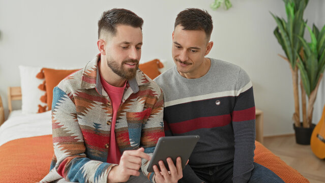 Two men on bed, one points finger to tablet while both lean in viewing the screen in bedroom; comfort connection.