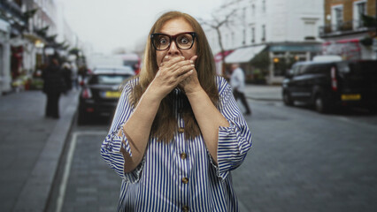 Fototapeta premium Senior woman wearing striped blouse and glasses covers her mouth with hands on a commercial city street beside parked cars and cafes; surprise caution.