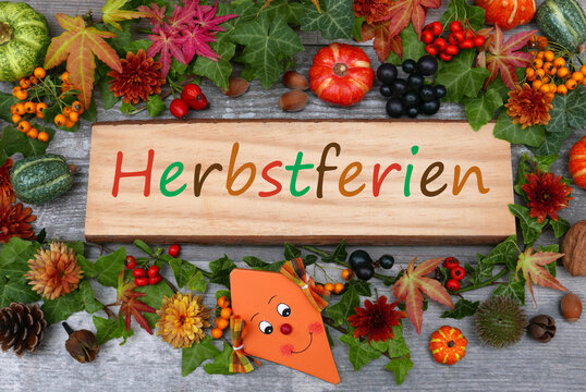 Holzschild mit dem bunten Schriftzug Herbstferien mit Naturdekoration, Zierk&uuml;rbissen, Bl&auml;ttern und einem l&auml;chelnden Papierdrachen