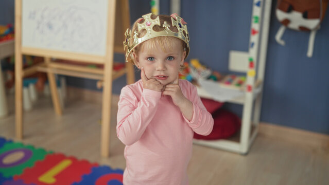 Little blonde girl adjusting a gold crown with hands in a preschool building, toys and easel nearby; playful imagination.