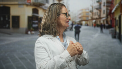 Fototapeta premium Hispanic woman doctor in white coat clasps hands to chest on a cobbled street with blurred storefronts and passersby; calm reflection.