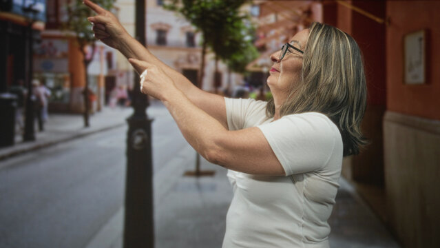 Grey haired woman points finger like a pretend gun on a city street in front of a building; playful gesture.