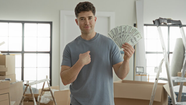 Man points to chest while holding a fan of dollars in a building amid moving boxes and ladder; confidence success.