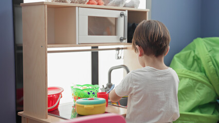 Naklejka premium Boy turns toy faucet at kitchen sink in building while playing with colorful pots, red cup and green basket on wooden play counter; childhood joy.