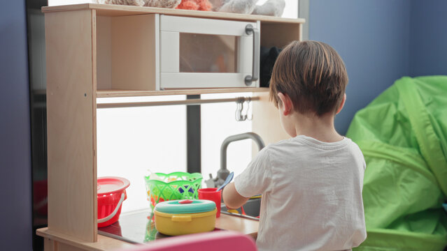 Boy turns toy faucet at kitchen sink in building while playing with colorful pots, red cup and green basket on wooden play counter; childhood joy.