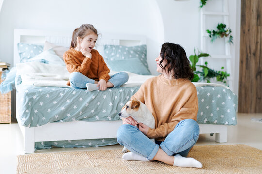 Cheerful little caucasian girl in an orange sweater talking to her young mother who holds a dog on the floor of a bedroom, ideal for family communication and parenting content