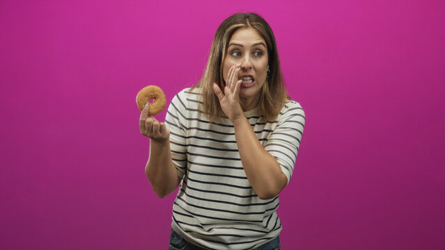 Woman holding donut and touching cheek with wry face in studio, striped shirt and visible hand gesture against pink backdrop; temptation curiosity.