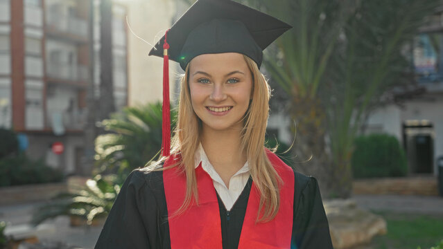 Woman graduate in cap and gown holding diploma and smiling in front of building; pride success celebration.