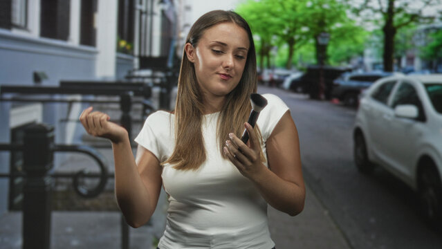 Woman holding makeup brush in hand on city street while tucking hair behind ear and glancing down; quiet contemplation.