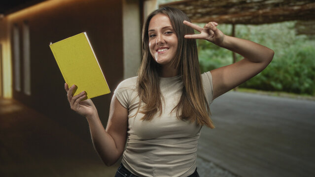 Woman holding yellow book and flashing peace sign in building, smiling and posing in a casual t shirt beneath wooden beams and greenery; playful curiosity.