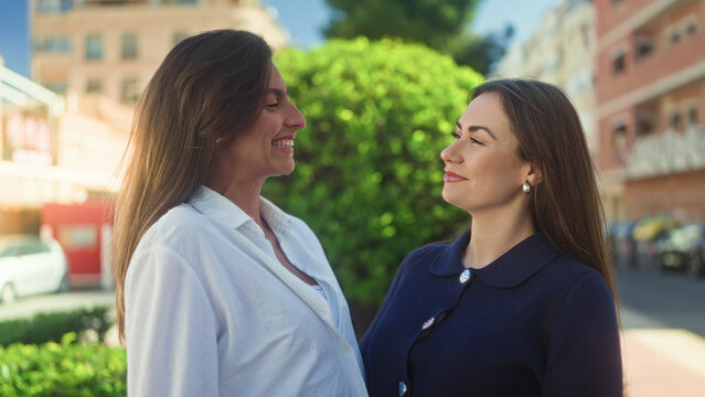 Two women smile with faces close and one arm around shoulder on a sunny street; warm friendship joy.