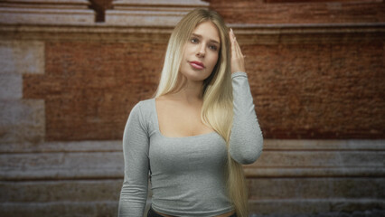 Fototapeta premium Woman pressing hand to forehead with closed eyes and long blonde hair standing in front of a brick building near stone steps; fatigue.