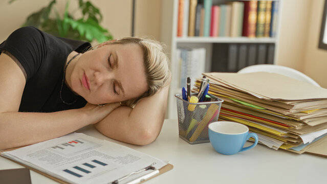 Woman resting head on desk beside computer, paperwork and coffee mug, arm folded and eyes closed in office building; exhaustion.