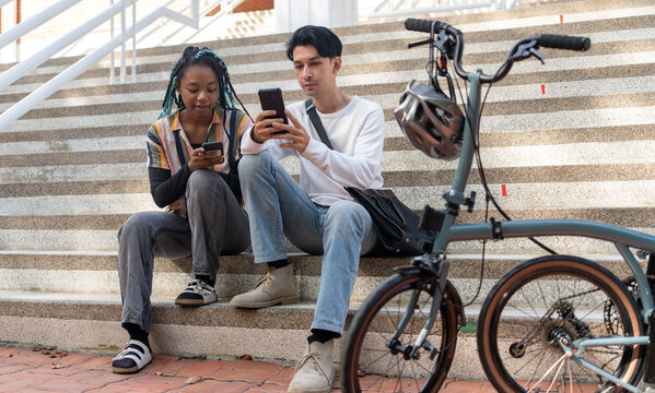Urban Commute: Two young adults, a man and a black woman, take a break on concrete stairs beside a foldable bicycle, engrossed in their smartphones. Bike to work.