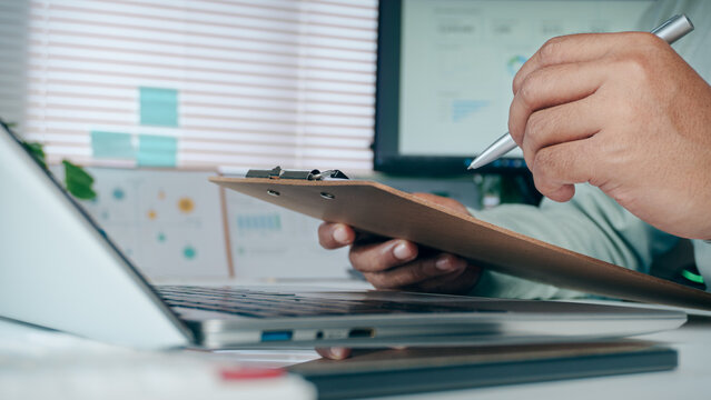 Business professional holding pen and clipboard with document report on office desk with laptop computer setup, concept of corporate management, paperwork review, and auditing.