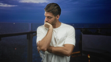 Fototapeta premium Man with beard in white shirt holding water bottle, hand on chin leaning on balcony railing of building overlooking ocean at dusk; quiet contemplation.