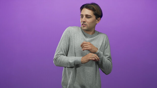Man in studio clutching his wrist and holding a hand to chest against a purple backdrop, wearing a gray sweater and ring; unease vulnerability.