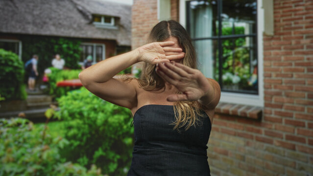 Woman shields eyes with hands and holds palms forward in front of brick building window and garden; shyness privacy solitude.