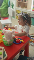 Little hispanic girl wearing a tiara at a red play table lifts a toy fork and reaches to feed a teddy in a building  playful imagination. © Krakenimages.com