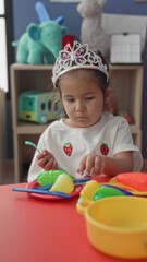 Girl in tiara holds toy fork and reaches with hand over colorful play dishes on red table inside building  quiet concentration. © Krakenimages.com