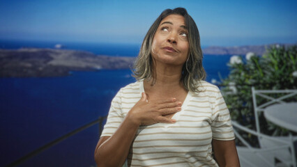 Fototapeta premium Woman middleaged hispanic with hand on chest looking up on building balcony with white railing, striped shirt and sea view; quiet gratitude.