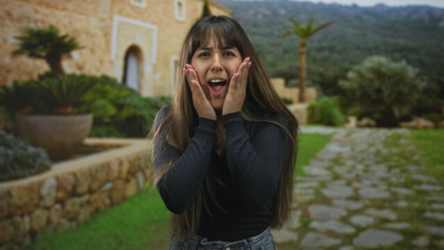 Young woman with hands on cheeks, open mouth and smile, touching her face in a stone courtyard by a villa entrance with potted plants and a cobblestone path; surprise moment.