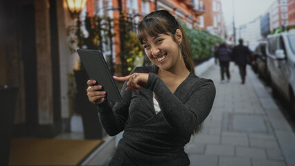 Young hispanic woman holding tablet points finger to screen on city street near storefront, smiling and engaging with device  joy engagement connection. © Krakenimages.com