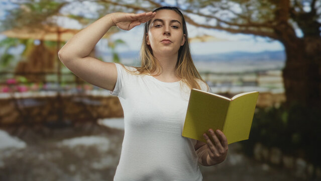 Woman holding yellow book salutes forehead while reading aloud beside a sunlit outdoor cafe patio in front of building; contemplation respect.
