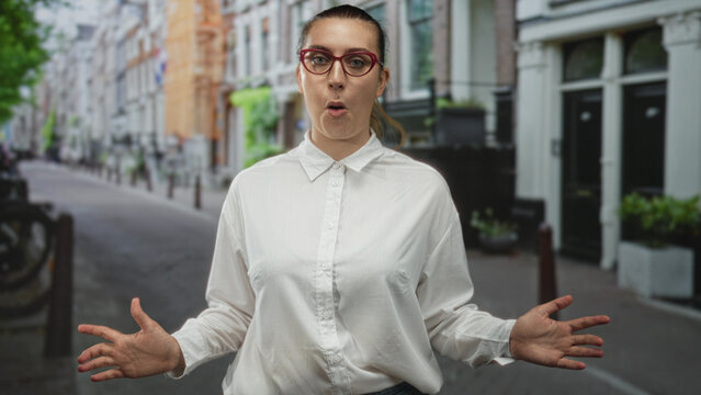 Woman gestures with open hands, wearing red glasses and white shirt, standing on a narrow city street lined with bicycles and townhouses; surprise conversation engagement.