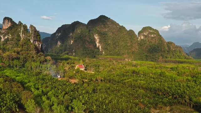 Drone flight over traditional Thai temple nestled among lush green jungle and towering limestone karst mountains in the scenic KHAO SOK National Park, Thailand. Bright sunset aerial view landscape