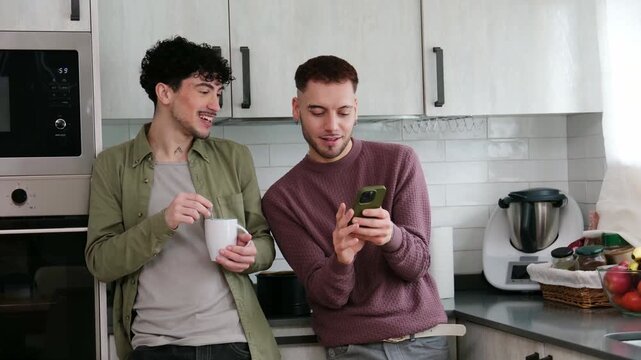Young male couple spending quality time together, talking and using a smartphone in their kitchen