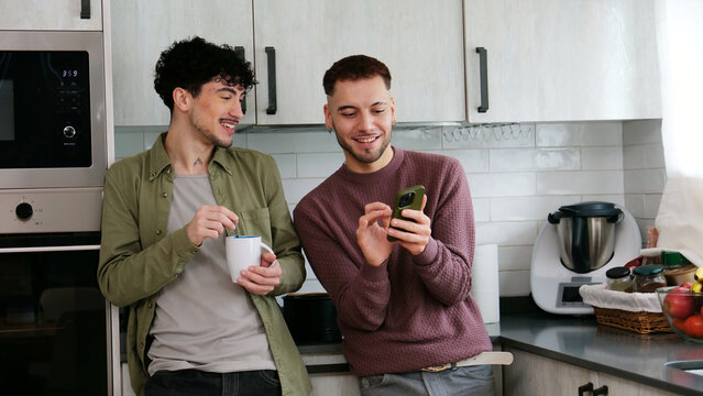 Happy gay couple smiling and interacting with smartphone in their modern kitchen