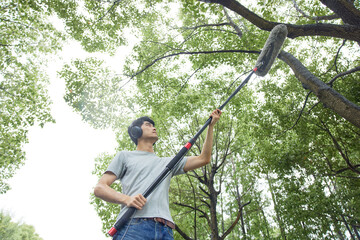 Young man recording audio outdoors with boom microphone under trees © HaXuanDieu