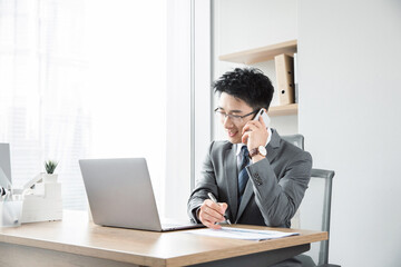 Fototapeta premium Young businessman talking on phone and taking notes at office desk