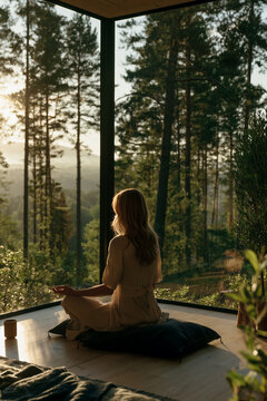 young woman meditating by large window overlooking forest, peaceful mindfulness meditation in scandinavian minimalist home, serene morning meditation with forest view in modern interior