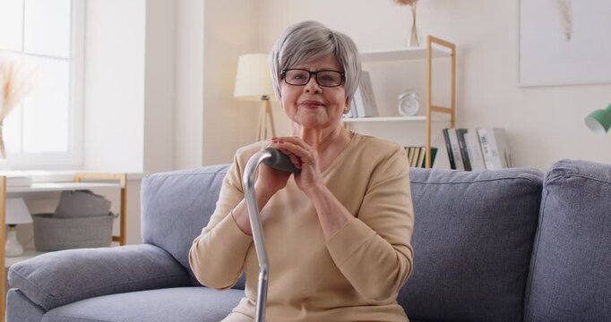 Elderly senior woman portrait with serious, thoughtful gaze holding crutch. Gray-haired smiling cheerful female sits on a sofa with a cane, wearing glasses, looking toward a window at home. 