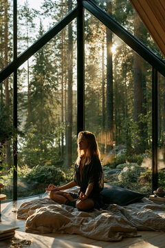 young woman meditating by large window overlooking forest, peaceful mindfulness meditation in scandinavian minimalist home, serene morning meditation with forest view in modern interior