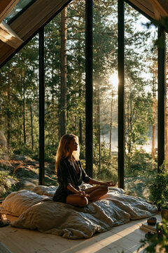 young woman meditating by large window overlooking forest, peaceful mindfulness meditation in scandinavian minimalist home, serene morning meditation with forest view in modern interior