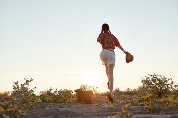 Fototapeta premium Young woman in a striped shirt and white shorts runs toward the setting sun on a dusty country trail, hat in hand, embodying carefree outdoor exploration.