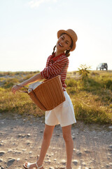 Fototapeta premium Young brunette woman in a striped red top and white shorts spins along a dusty country road with a wicker basket and straw hat, enjoying a sunny carefree moment.
