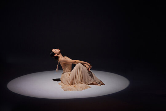 Graceful female dancer sitting in spotlight on dark stage
