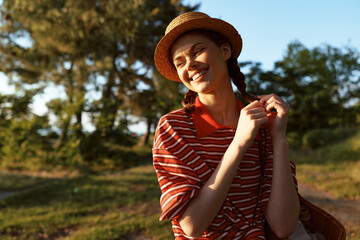 Naklejka premium Young woman in a striped red and white shirt smiles in bright sunlight in a park setting. Outdoor lifestyle portrait with natural light, casual mood, warm tones.