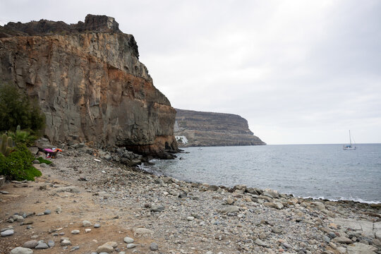 Imposante Steilklippe an einem steinigen K&uuml;stenstreifen mit Blick auf das ruhige Meer und ein entferntes Segelboot unter bedecktem Himmel. Eine raue und urspr&uuml;ngliche Landschaft voller Natur.