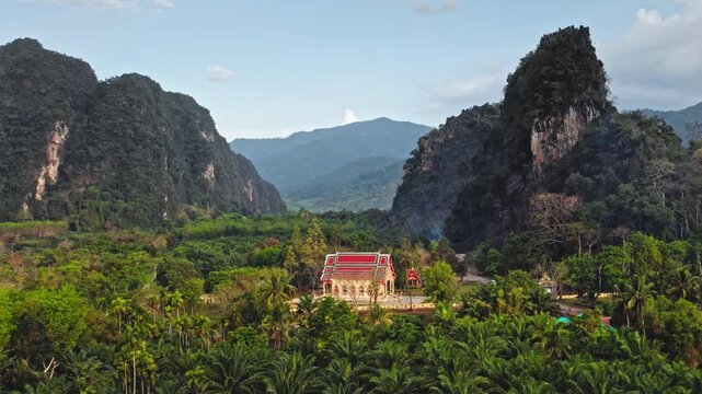 Buddhist temple nestled in a lush green jungle valley surrounded by dramatic limestone cliffs. Serene and spiritual landscape of Khao Sok National Park, Thailand. Aerial view drone flight zoom out