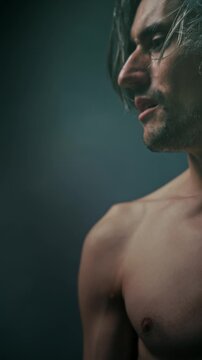 humility acceptance Close-up side-profile portrait of a young adult man with shoulder-length wavy hair and bare shoulders, serious expression against a dark blurred background with soft lighting