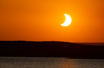 Bright crescent solar eclipse in vivid orange sky over San Salvador reservoir, Spain. © PABLO