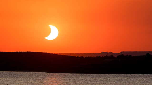 Crescent solar eclipse setting in dramatic red-orange sky over San Salvador reservoir, Spain.
